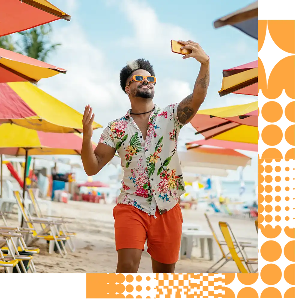 Man on a beach wearing a floral shirt taking a selfie.